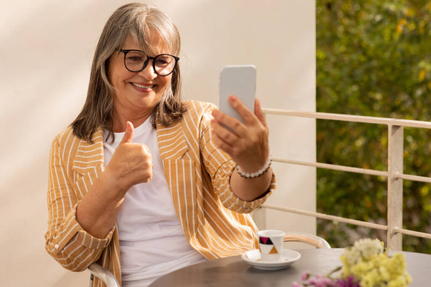 Szczęśliwa, biała, siwa kobieta w okularach siedzi przy stole w kawiarni, robi selfie, ma rozmowę wideo na smartfonie, pokazuje gest kciuka w górze, na świeżym powietrzu. Pani lubi aplikację, odpoczynek, komunikację zdalnie - Zdjęcie, obraz