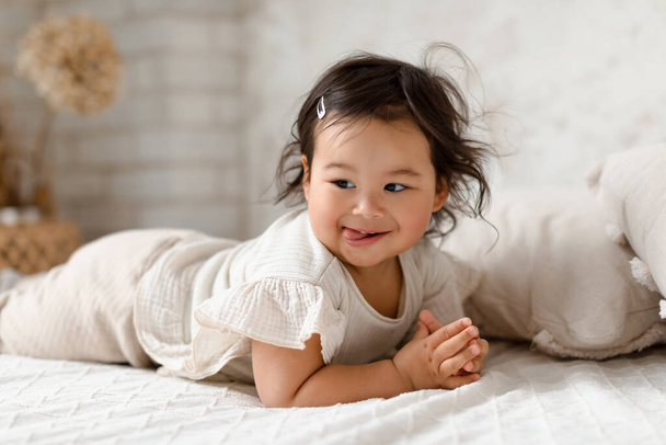 Дитинство. Мила корейська дівчинка позує, дивлячись в сторону брехні на stomach на ліжко в затишний домашній інтер'єр. Знімок смішного маленького Тоддлера в зручному одязі Розважаючись в спальні
 - Фото, зображення