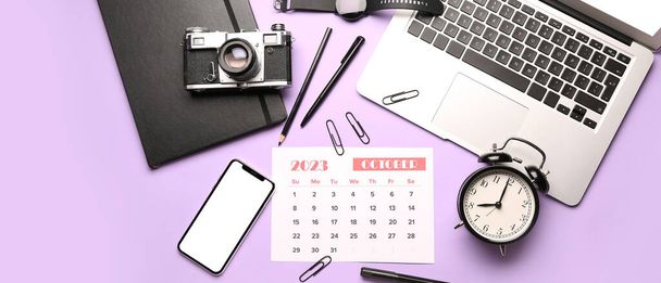 Kalender für Oktober 2023, Wecker, Laptop, Handy, Fotokamera und Bürobedarf auf fliederfarbenem Hintergrund - Foto, Bild