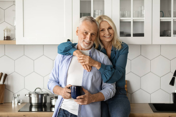 Портрет любящей пожилой пары, расслабляющей себя с кофе в интерьере кухни, романтические пожилые супруги, обнимающиеся и улыбающиеся перед камерой, зрелая жена и муж, сближающиеся дома, копировальное пространство - Фото, изображение