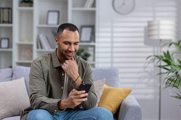 Smiling young hispanic man reading message, news from phone. Sitting on the sofa at home, talking on a video call. - Foto, Imagen