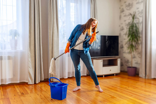 Αστεία χαρούμενη νεαρή γυναίκα με σφουγγαρίστρα να τραγουδάει, να χορεύει και να διασκεδάζει καθαρίζοντας το πάτωμα. Η νοικοκυρά απολαμβάνει τις δουλειές του σπιτιού, καθαρίζει το σπίτι δημιουργικά. - Φωτογραφία, εικόνα