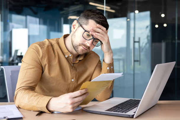 Shocked young man businessman sits in the office at the table and reads the received letter, documents. He holds his head worriedly. - Photo, Image