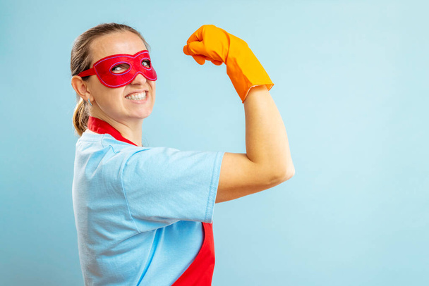 Confidence superhero house cleaner showing her powerful muscle arm with wearing red apron and eye mask on blue background - Photo, Image