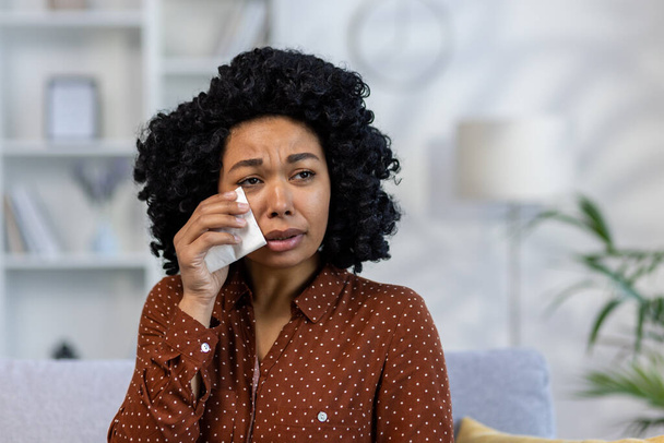 Close-up photo. Portrait of upset young African American woman sitting on sofa at home and crying, suffering from depression, wiping tears with napkin. - Foto, immagini