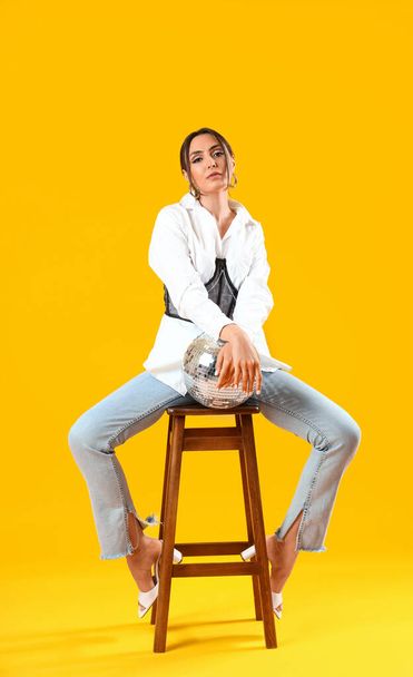Elegante bella donna con palla da discoteca seduta sulla sedia e posa sullo sfondo giallo - Foto, immagini