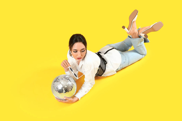 Красивая молодая женщина в стильных солнцезащитных очках и диско мяч лежит на желтом фоне - Фото, изображение