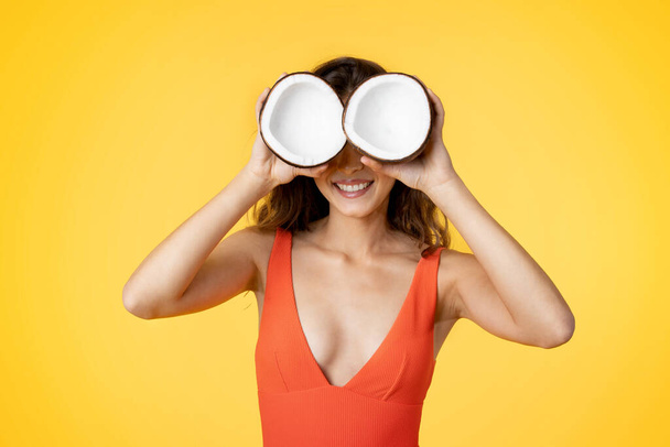 Cheerful millennial mixed race lady in swimsuit puts coconuts to eyes, has fun, enjoy vacation, isolated on yellow studio background. Good holiday trip in summer, weekend, free time, ad and offer - Foto, imagen