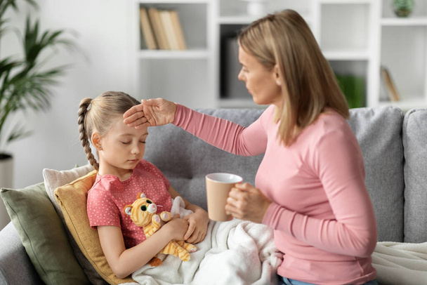 Opiekuńcza młoda matka dając gorącą herbatę do chorej małej córeczki i dotykając jej czoła, kochający mama kaukaska dbanie o chorych kobiet w domu, słodkie dziewczyny cierpiące na grypę sezonową lub zimno - Zdjęcie, obraz
