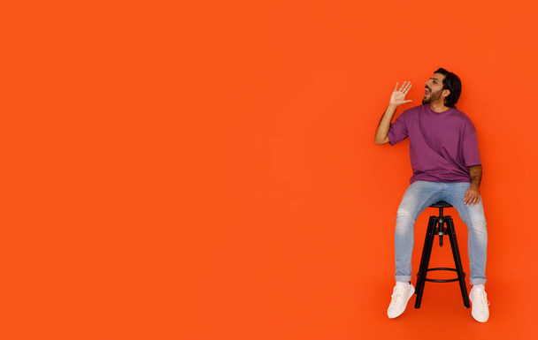 Cool heureux beau jeune homme oriental portant tenue décontractée élégant assis sur la chaise sur fond de studio orange, criant vers l'espace de copie pour la publicité, faire une annonce, plan complet - Photo, image