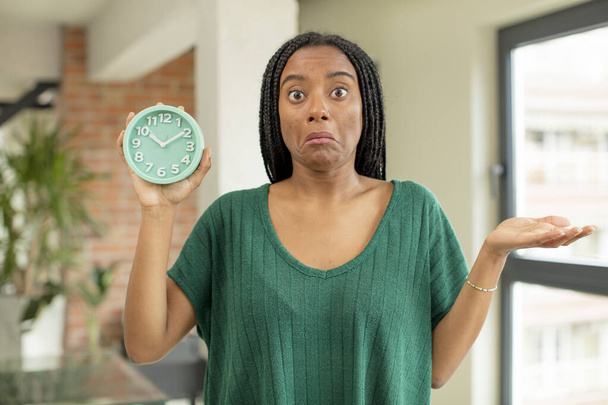 black afro woman shrugging, feeling confused and uncertain. alarm clock concept - Foto, Imagen