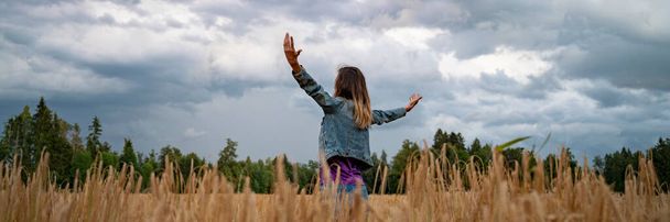 Wide view image of a young woman celebrating and enjoying life standing with her arms spread widely in the middle of golden wheat field under a dramatic cloudy summer sky. - Photo, Image