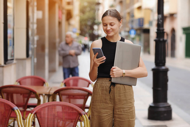 Fiatal elegáns üzletasszony szabadúszó tartja laptop és okostelefon az európai város utcájában. Fogalom a távoli munka vagy szabadidő a kütyü a szabadban. A modern kommunikáció gondolata. - Fotó, kép
