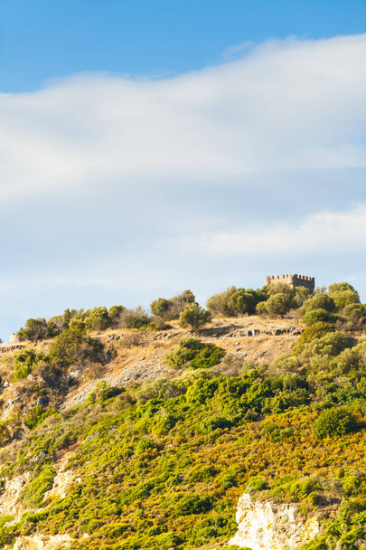 Greek idyllic hills with small shrubs of Mediterranean flora and little castle against cloudy sky. - Foto, imagen