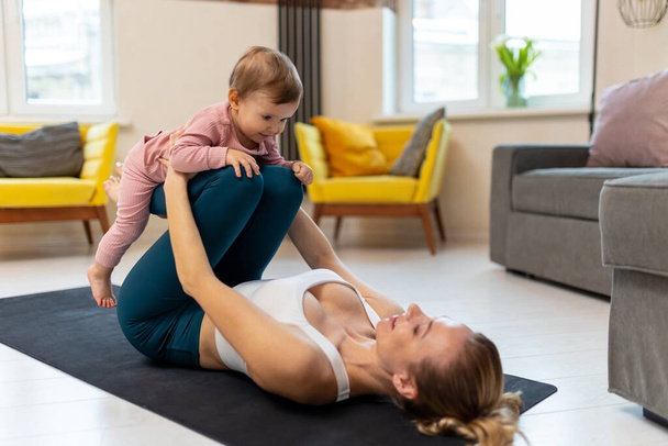 Young sporty mother in sportswear and baby girl working out exercising together at home in living room. - Fotó, kép