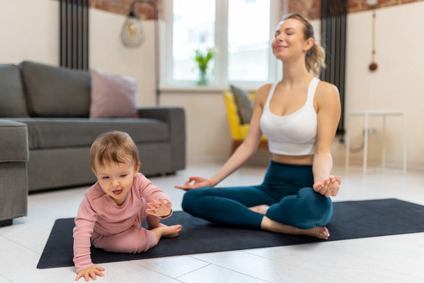Athletic woman in top and leggins practicing yoga with her baby daughter in living room. - Foto, Imagem