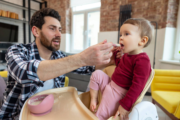 Lure first meal. Dad feeding toddler child with spoon for lunch in modern home interior. - Foto, immagini