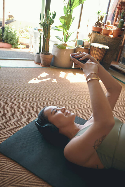 Aplicativos de fitness tornaram-se seu treinador virtual. uma jovem mulher usando fones de ouvido e usando um celular enquanto estava deitada em um tapete de exercícios em casa - Foto, Imagem