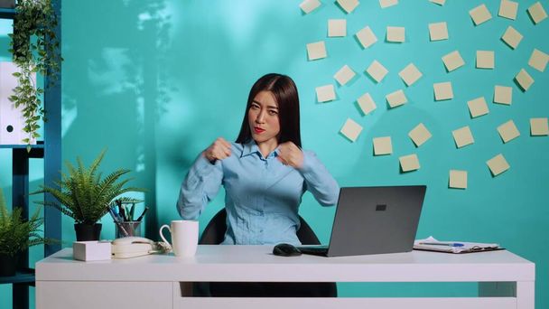 Happy playful asian office worker having fun pretending to fight, throwing punches. Funny high spirited businesswoman mock boxing in relaxed colourful workplace over blue studio background - Photo, Image