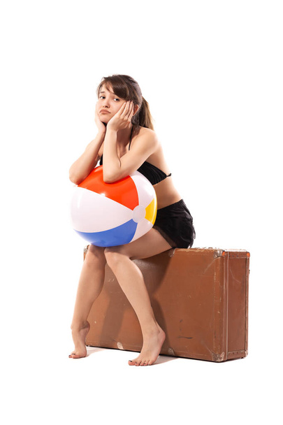Sad girl in bikini and black dress with colorful inflatable beach ball sitting on big vintage bag. Summer travel concept. - Фото, изображение