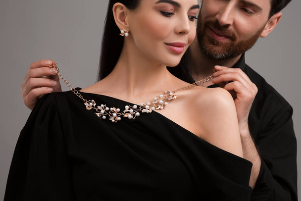 Мужчина надевает элегантное ожерелье на красивую женщину на сером фоне, крупным планом - Фото, изображение