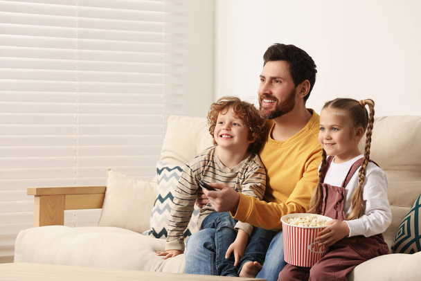 Pai feliz e crianças assistindo TV com pipocas no sofá dentro de casa, espaço para texto - Foto, Imagem