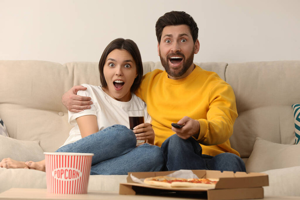 Surprised couple watching TV with popcorn and pizza on sofa at home - Foto, imagen