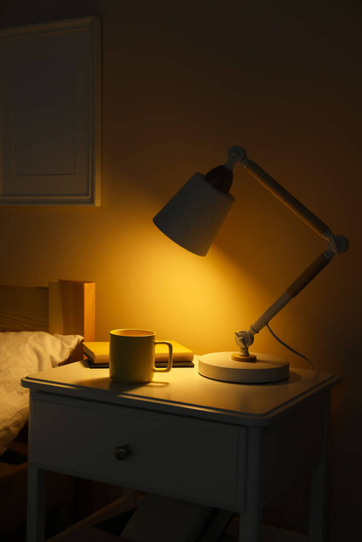 Стильна сучасна настільна лампа, книги та чашка напою на білій тумбочці в темній спальні
 - Фото, зображення
