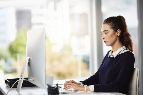 Почти закончил с этим заданием. привлекательная молодая деловая женщина, сидящая за своим столом и пользующаяся компьютером в современном офисе - Фото, изображение