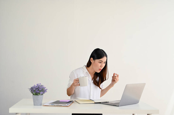 Eine erstaunt und überrascht wirkende junge asiatische Mitarbeiterin blickt auf ihren Laptop und liest eine unerwartete E-Mail, während sie an ihrem Schreibtisch sitzt. isolierte weiße Studiohintergrund - Foto, Bild