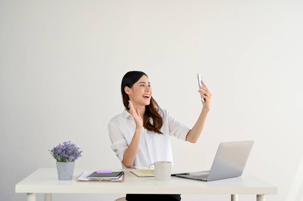 Egy elbűvölő és vidám fiatal ázsiai üzletasszony vagy női irodai dolgozó beszélget a barátaival egy videóhíváson keresztül az íróasztalánál lévő okostelefonján. izolált fehér háttér - Fotó, kép