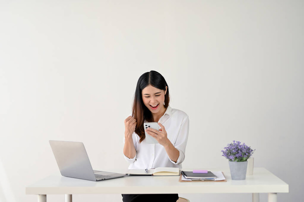 Heyecanlı ve neşeli Asyalı genç iş kadını telefon ekranına bakıyor ve masasında otururken yumruğunu gösteriyor. Kutlama, sevinç, iyi haber, kazanan piyango, soyutlanmış beyaz arka plan - Fotoğraf, Görsel