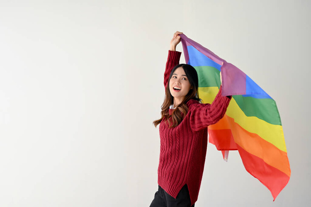 Счастливая и красивая молодая азиатка в повседневной одежде с радужным ЛГБТ-флагом стоит на изолированном белом фоне студии. ЛГБТК + месяц гордости - Фото, изображение