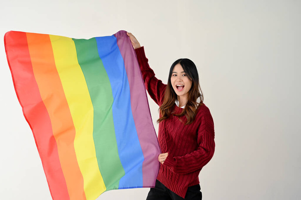 Une jeune Asiatique heureuse en tenue décontractée avec un drapeau LGBT arc-en-ciel se dresse sur un fond de studio blanc isolé. Mois de la fierté LGBTQ +, défilé LGBTQ - Photo, image