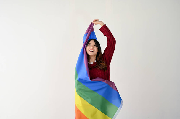 Šťastná mladá Asiatka v neformálním oblečení s duhovou LGBT vlajkou stojí nad izolovaným bílým studiem pozadí. LGBTQ + měsíc hrdosti - Fotografie, Obrázek