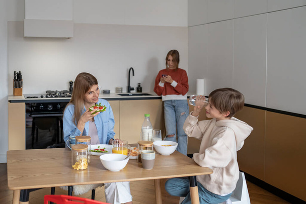 Family breakfast mom and kids. Young woman, teenager son eat at table in kitchen and talk. Eldest daughter stands aside and chats on phone in secret from mother. Relationship children and parents - Photo, Image