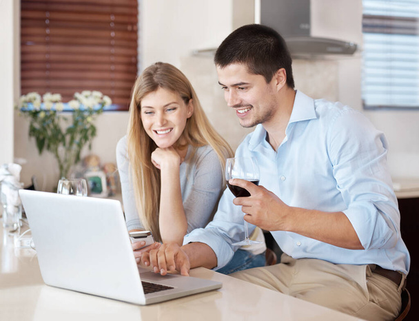 Entspannen und gemeinsam im Netz surfen. Ein glückliches Paar genießt ein Glas Wein in der Küche und schaut gemeinsam auf einen Laptop - Foto, Bild