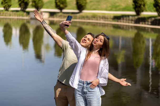Portrait of smiling couple drinking takeaway coffee from paper cups and taking selfie photo on cellphone while walking in spring park - Photo, Image