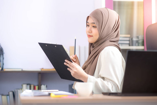 L'impiegato, una donna musulmana asiatica, è seduta davanti al computer portatile alla scrivania e sta guardando i file con successo in ufficio.. - Foto, immagini