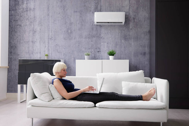 Young Happy Woman Lying Under Air Conditioner On Couch Using Laptop At Home - Φωτογραφία, εικόνα