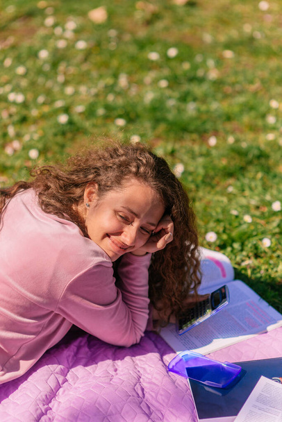 Кудрявая девушка лежит на фиолетовом одеяле в парке, разговаривает со своей подругой и улыбается - Фото, изображение