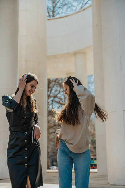 Two girls looking down while dancing. They are listening to music on their headphones while having fun in the city - Foto, Imagen