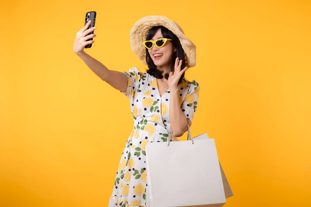 Portrait attrayant jeune femme asiatique heureuse en utilisant un smartphone avec robe de printemps à la mode, chapeau, lunettes de soleil mode et sac en papier isolé sur fond jaune. Vente d'été concept de shopping. - Photo, image