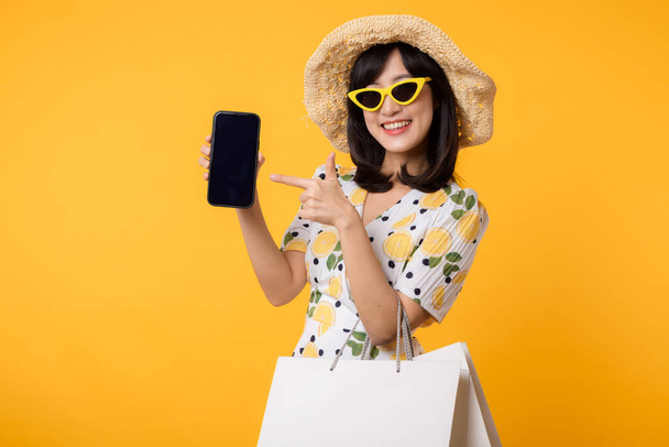 Portrait happy young asian woman showing blank display smartphone with trendy springtime dress, hat, sunglasses fashion and paper bag isolated on yellow background. Summertime sale shopping concept. - Foto, afbeelding