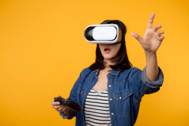 アジアの若い女性は、仮想現実ヘッドセットゲームエンターテイメントを着用し、黄色の背景に隔離されたジョイスティックコントローラを保持幸せな成功。サイバー未来のビデオゲームのコンセプトの勝者. - 写真・画像