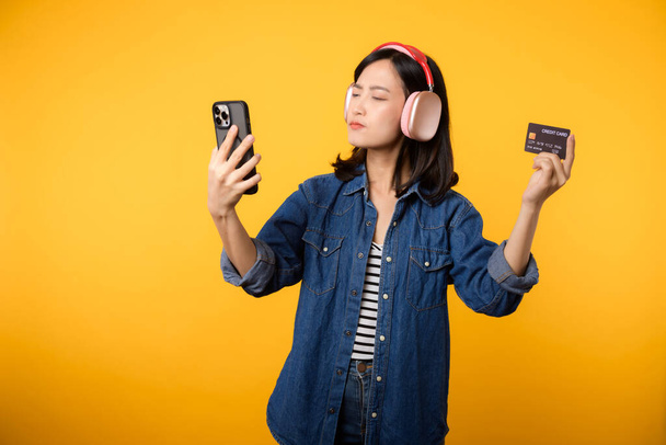 Muotokuva aasialainen nuori nainen epäilevästi kasvot yllään denim vaatteita ja kuulokkeet tilalla älypuhelin ja luottokortti. Ostoksia verkossa matkapuhelin viihde elämäntapa käsite. - Valokuva, kuva