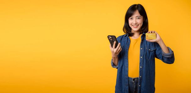 Retrato hermosa asiática joven sonrisa feliz vestido con chaqueta de mezclilla que muestra teléfono inteligente y tarjeta de crédito aislado en el fondo del estudio amarillo. Compras en línea smartphone concepto de aplicación - Foto, imagen