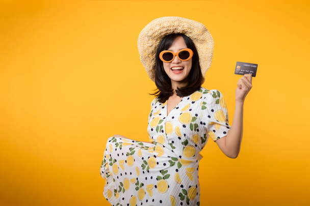 Sarı arka planda plastik kredi kartını gösteren bahar elbisesi içinde gülümseyen Asyalı genç bir kadın portresi. Ödeme, para ve alışveriş ödeme kavramı. - Fotoğraf, Görsel