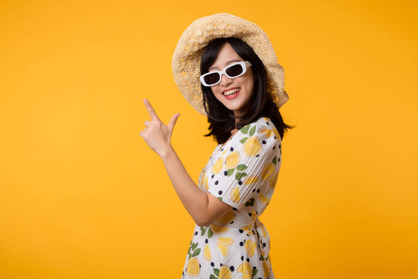 Portret młodej pięknej azjatyckiej kobiety noszącej sukienkę wiosną i wskazującej na miejsce do kopiowania reklam odizolowanych na żółtym tle. Koncepcja banera reklamowego Summertime. - Zdjęcie, obraz