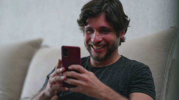 Joyful young man liking content online while holding smartphone device in hand. Male person in 30s excited about entertainment media sitting on couch - Foto, imagen